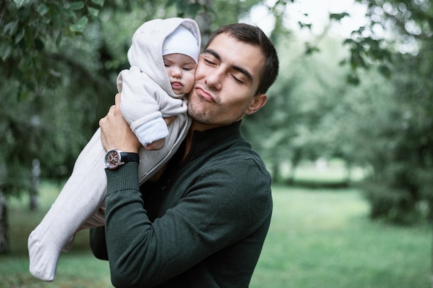 Papá de hombre de pelo oscuro en chaqueta verde con bebé en sus manos en el parque, concepto de paternidad