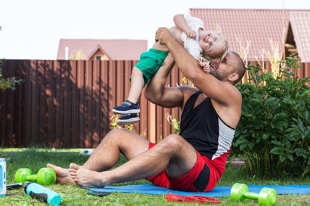 Papá joven atleta con su pequeño hijo alegre practica deportes, estirándose en la colchoneta en un día caluroso en el jardín cerca de la casa