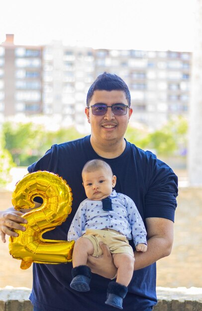 Foto papá joven con un bebé en brazos en un parque y un globo número dos