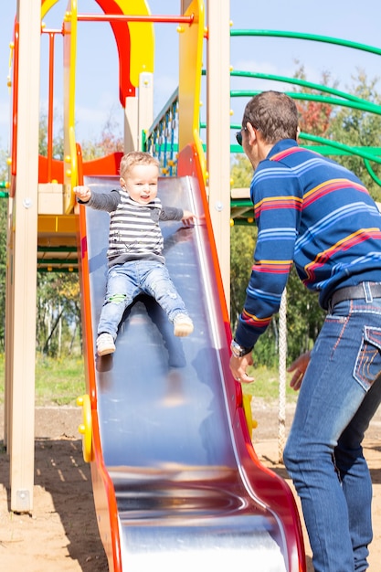 Papá juega con un niño en el patio de recreo. El niño se sale del tobogán