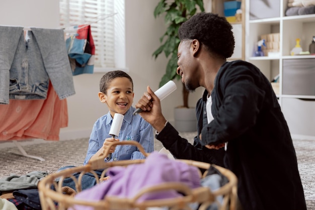 Papá jugando con su hijo mientras limpia haciendo las tareas domésticas Hombre cantando para limpiar el rollo de ropa sacada de la lavadora seca para clasificar