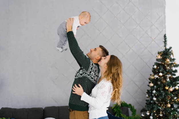 Papá levantó a su pequeño hijo en sus brazos, mamá abraza a papá y miran a su bebé juntos cerca del árbol de Navidad.