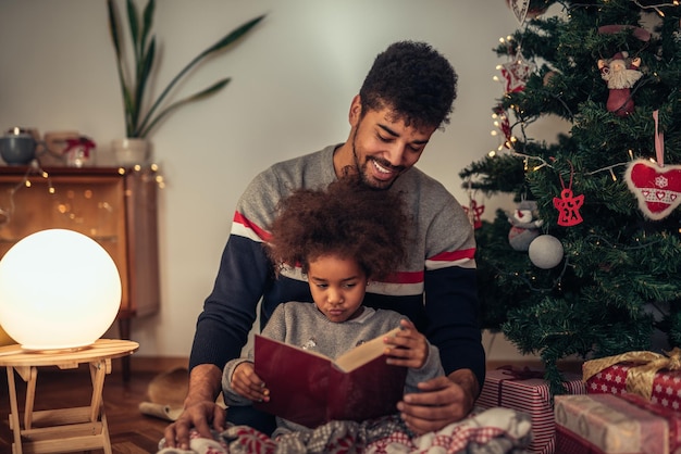 Papa liest seiner Tochter Märchen vor, während er neben dem Weihnachtsbaum sitzt