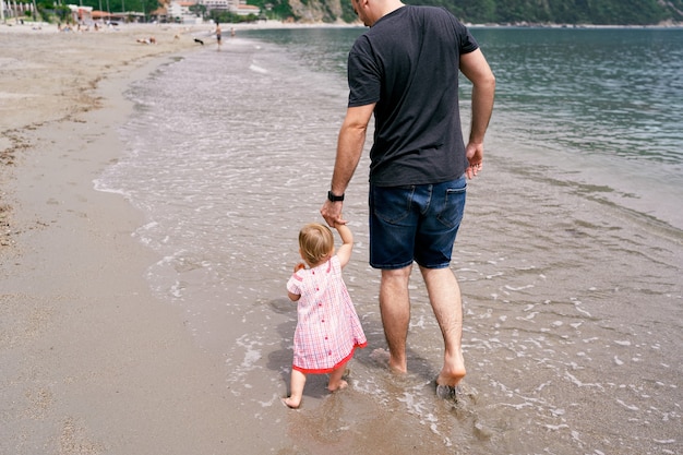 Papá lleva a una niña de la mano a lo largo de la orilla arenosa