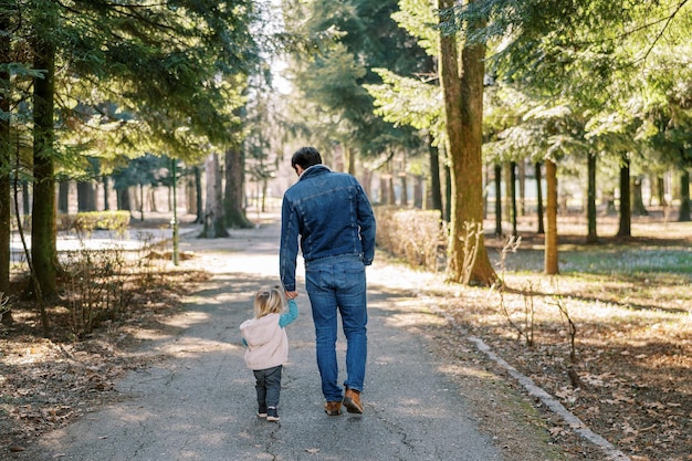 Foto papá lleva a una niña por la mano a través de un parque de primavera soleado vista trasera