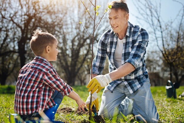 Papá maduro enseñando a su hijo pequeño cómo cuidar la naturaleza colocando un árbol en el patio de una casa de campo