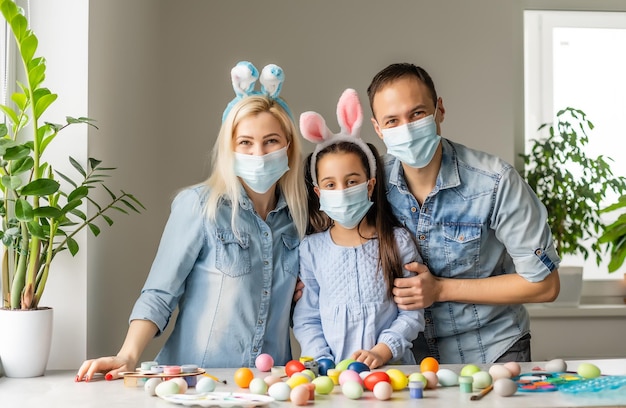 Papá, mamá e hija en la mesa con máscaras médicas están pintando huevos de Pascua para las vacaciones. Feliz pascua familia coronavirus en cuarentena. Feliz en casa.