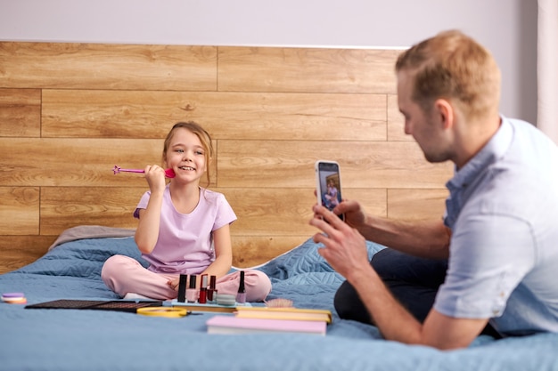 Papá moderno toma una foto de su hija de moda usando cosméticos, el hombre y el niño se sientan juntos en la cama