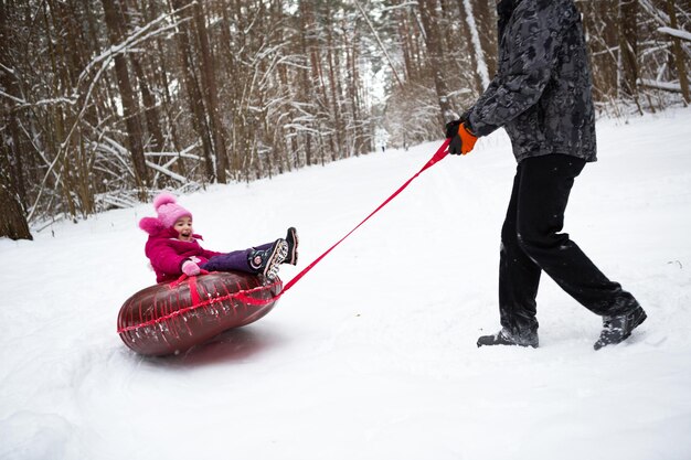 Papá monta a su hija en un tubo de un tobogán en invierno en una nevada. Tubing, deportes de invierno al aire libre y entretenimiento familiar. Ropa de abrigo, gorro de punto con pompón. Ayuda y apoyo, valores familiares