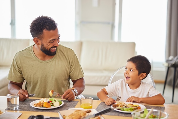 Papá y niño felices en la mesa para el almuerzo comida saludable y unión en la sala de estar padre niño y niño y sonrisa en casa para la nutrición tiempo familiar y comida del domingo con historia divertida o bromeando juntos