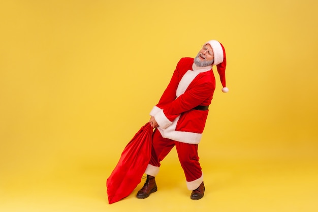 Papá Noel arrastrando su bolsa grande y pesada llena de regalos, con el ceño fruncido, tratando de levantarla.