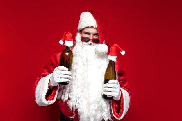 Papá Noel borracho con sombrero y copas festivas bebe vino de una botella de fondo rojo