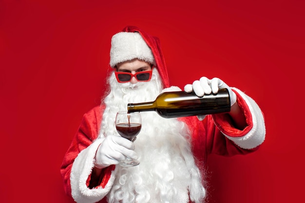 Foto papá noel borracho con sombrero y copas festivas bebe vino de una botella de fondo rojo