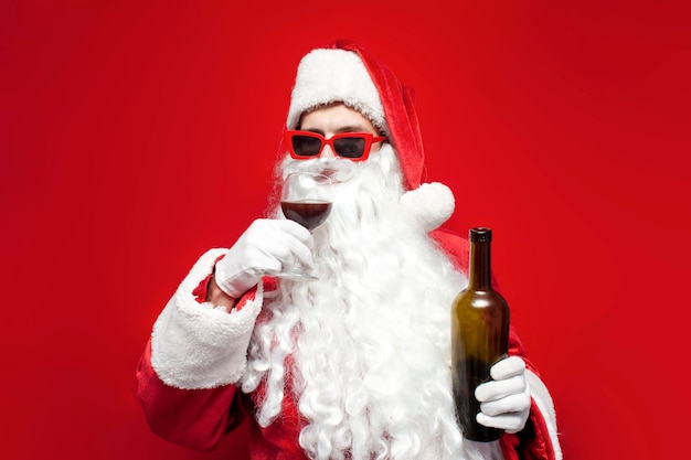 Papá Noel borracho con sombrero y copas festivas bebe vino de cristal sobre fondo rojo