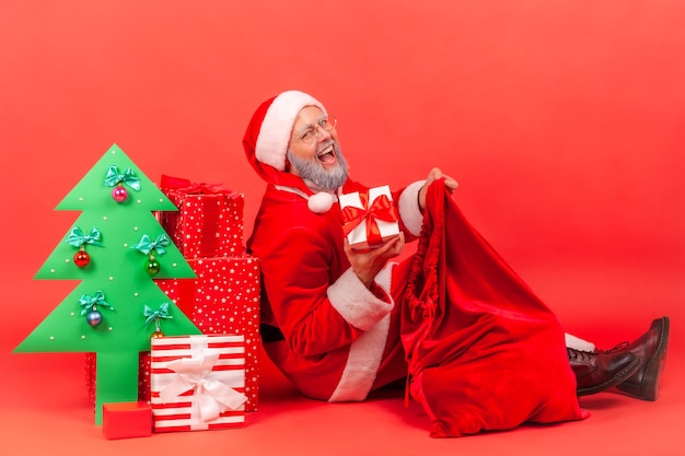 Papá Noel con humor festivo, sacando la caja presente de la bolsa, mirando a cámara y sonriendo.