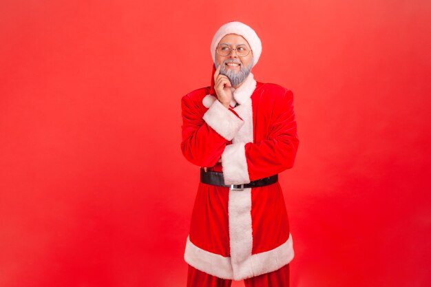 Papá Noel de pie con el dedo en la mejilla soñando y pensando en recuerdos agradables.