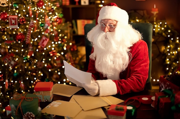 Papá Noel prepara regalos navideños para niños en su escritorio en casa mientras lee listas de deseos