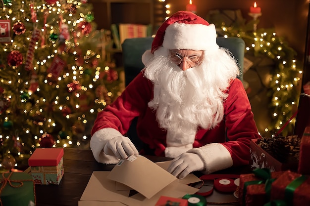 Papá Noel prepara regalos para niños para Navidad en su escritorio en casa mientras lee cartas