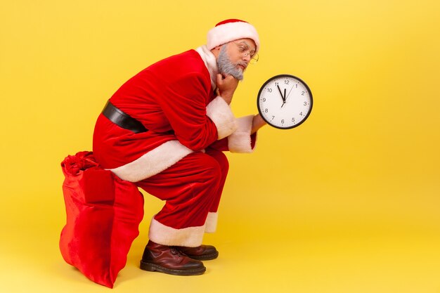 Papá Noel sentado en una gran bolsa roja con regalos y reloj de pared, tiempo de espera para dar presente.