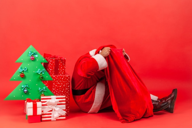 Papá Noel sentado en el piso cerca del árbol de Navidad de papel decorativo y regalos, poniendo la cabeza en bolsas