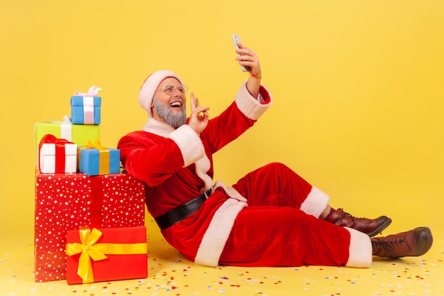 Papá Noel sentado en el piso cerca de regalos, con videollamada, mostrando el signo v a los seguidores.