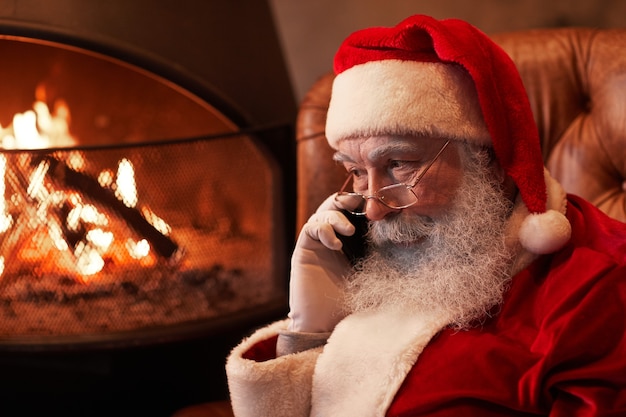 Papá Noel serio en anteojos sentado en la chimenea y hablando por teléfono móvil en una habitación oscura