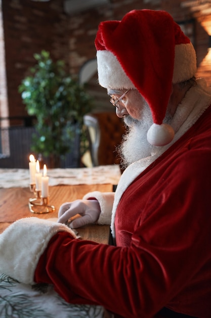 Papá Noel serio con gorra roja y vasos sentado en la mesa de madera con velas encendidas y leyendo ...