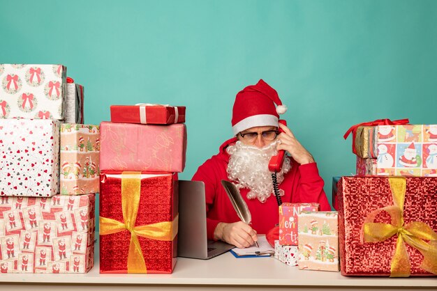 Papá Noel con sombrero trabaja en su oficina con regalos, preparándose para las vacaciones.