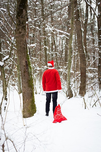 Un Papá Noel tradicional con un sombrero y una bolsa de regalos camina por el bosque llevando regalos. Vista trasera.