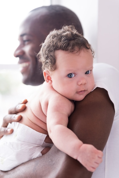 Papá orgulloso y feliz. Feliz joven africano sosteniendo a su pequeño bebé y sonriendo mientras está de pie cerca de la ventana