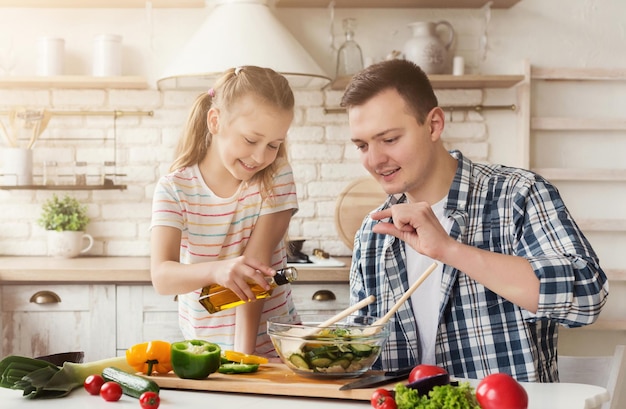 Papá positivo e hija pequeña cocinando en la cocina de casa. Hombre enseñando a la niña a agregar la cantidad correcta de aderezo para ensaladas, espacio de copia