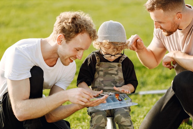 Papa, sein Freund und sein kleiner Junge verbringen gemeinsam Zeit im Freien. Gelockter Kleinkindjunge, der einen khakifarbenen Overall trägt. Junge, der richtig fischt