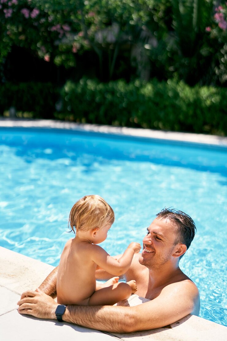 El papá sonriente está de pie en el agua y abraza a un niño pequeño sentado en el borde de la piscina