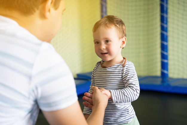 Papá sostiene la mano de un niño que salta en un trampolín Tiempo en familia Niño de un año y medio Entretenimiento activo Niño pequeño Enfoque en los ojos del niño