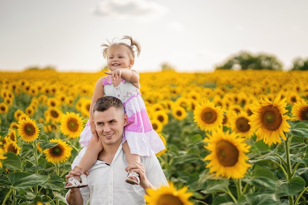 Foto papá sostiene sobre sus hombros a una pequeña hija. la niña señala algo con su dedo. campo de girasol. puesta de sol