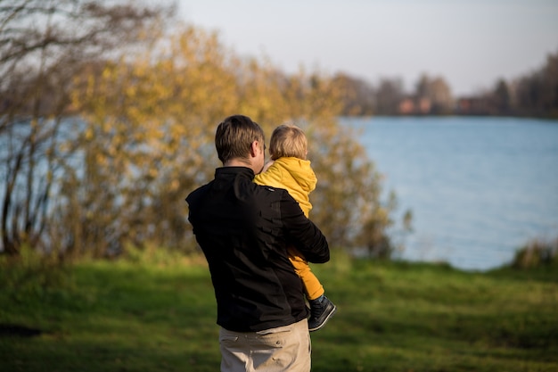 Papá sostiene a su hijo con una chaqueta amarilla en sus brazos y mira el lago