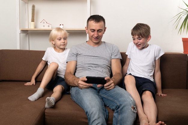Papá y sus dos hijos están sentados en el sofá y miran un video por teléfono. Familia feliz.