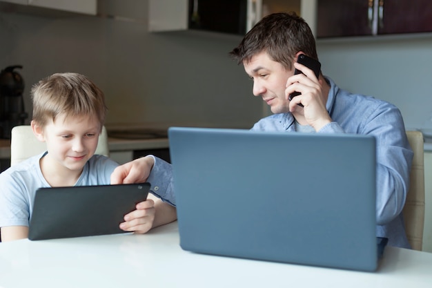 Papá trabaja desde su casa en una computadora portátil y su hijo estudia desde su casa en una tableta