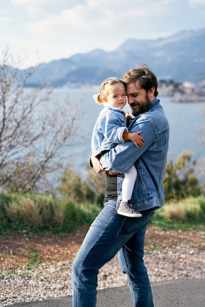 Papai carrega uma menina nos braços caminhando pela estrada tendo como pano de fundo o mar