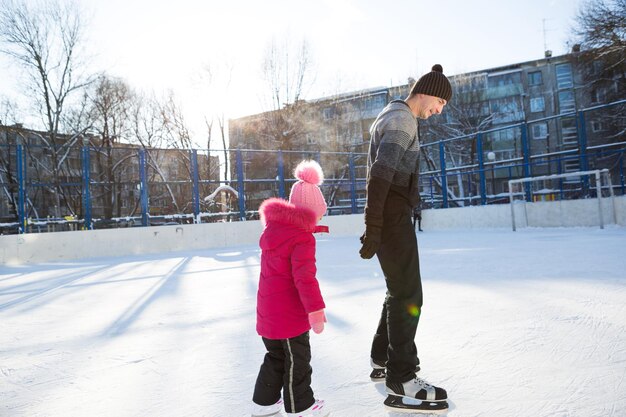 Papai ensina sua filha a patinar no gelo em um rinque de patinação no pátio de prédios de vários andares da cidade. dia gelado de sol de inverno, esportes de inverno ativos e estilo de vida