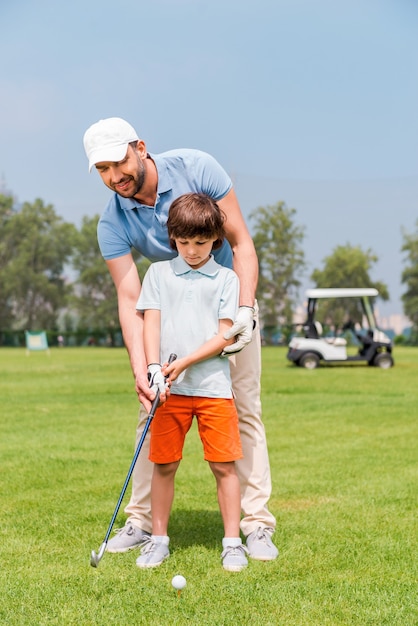 Papai está sempre perto para me ajudar. jovem alegre ensinando seu filho a jogar golfe enquanto está no campo de golfe