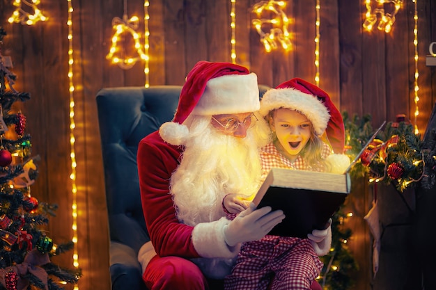 Papai Noel abre e lê um livro de mágica com uma garotinha linda e espantada de pijama