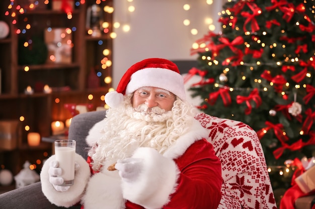 Papai Noel autêntico com biscoito e copo de leite sentado na poltrona em um quarto decorado para o Natal