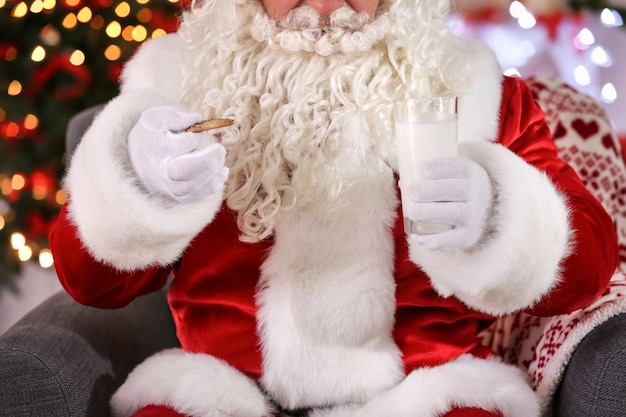 Papai Noel autêntico segurando um copo de leite e biscoito contra as luzes de Natal desfocadas