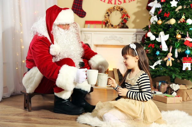 Papai Noel bebendo chocolate quente com uma linda garotinha perto da árvore de Natal em casa