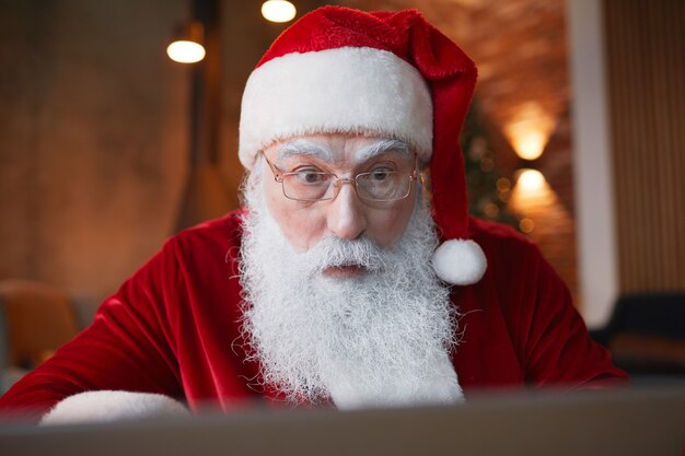 Papai Noel bonito chocado com barba branca usando laptop enquanto lê notícias da internet