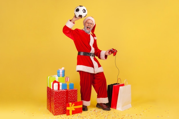 Papai noel com bola de futebol, segurando o joystick, comemorando a vitória no videogame de futebol.