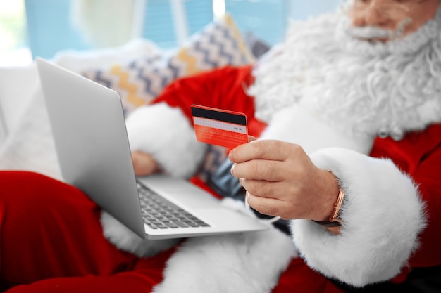 Papai Noel com cartão de crédito e laptop no sofá em casa
