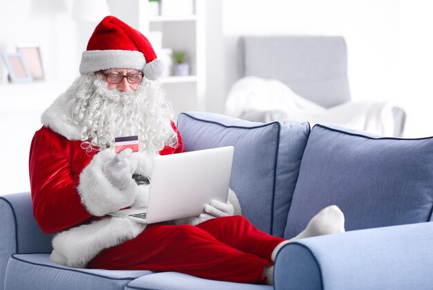 Papai Noel com laptop e cartão de crédito no sofá em casa