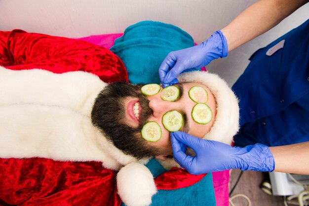 Papai Noel com máscara de pepino em spa de beleza. Homem de Natal com roupas de Papai Noel em spa de beleza.